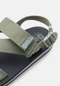 Green fabric sandals with adjustable straps, a textured dark blue sole, and branded accent. Includes a plastic buckle and woven label.