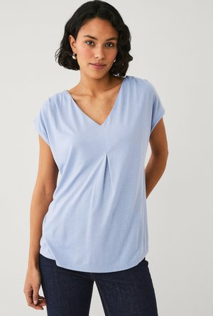 Kvinde med kort mørkt hår iført lyseblå V-hals bluse og mørke jeans, stående mod en ensfarvet lys baggrund.