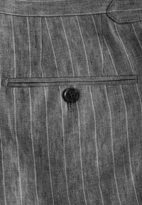 Tissu en lin gris avec de fines rayures blanches, comportant une poche avec un bouton noir à quatre trous au milieu.