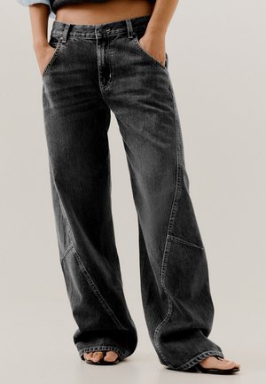 Personne portant un jean noir ample et délavé, les mains dans les poches, et des sandales noires à bouts ouverts sur un fond uni.