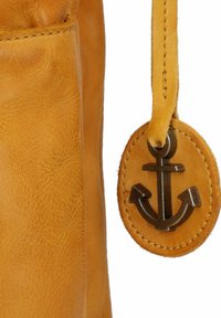 Sezione di borsa in pelle gialla con texture visibile, con un'etichetta rotonda che espone un emblema di ancora metallica attaccata da una tracolla in pelle.