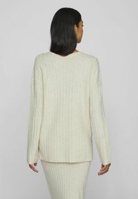Suéter de punto acanalado en beige, con escote en V, mangas largas y un dobladillo trasero ligeramente más largo, hecho de una tela suave y texturizada.