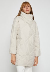 Beige quiltet puffjakke med stor krave, trykknaplukninger og skrå syninger. Har afslappede ærmer og en lang facon.