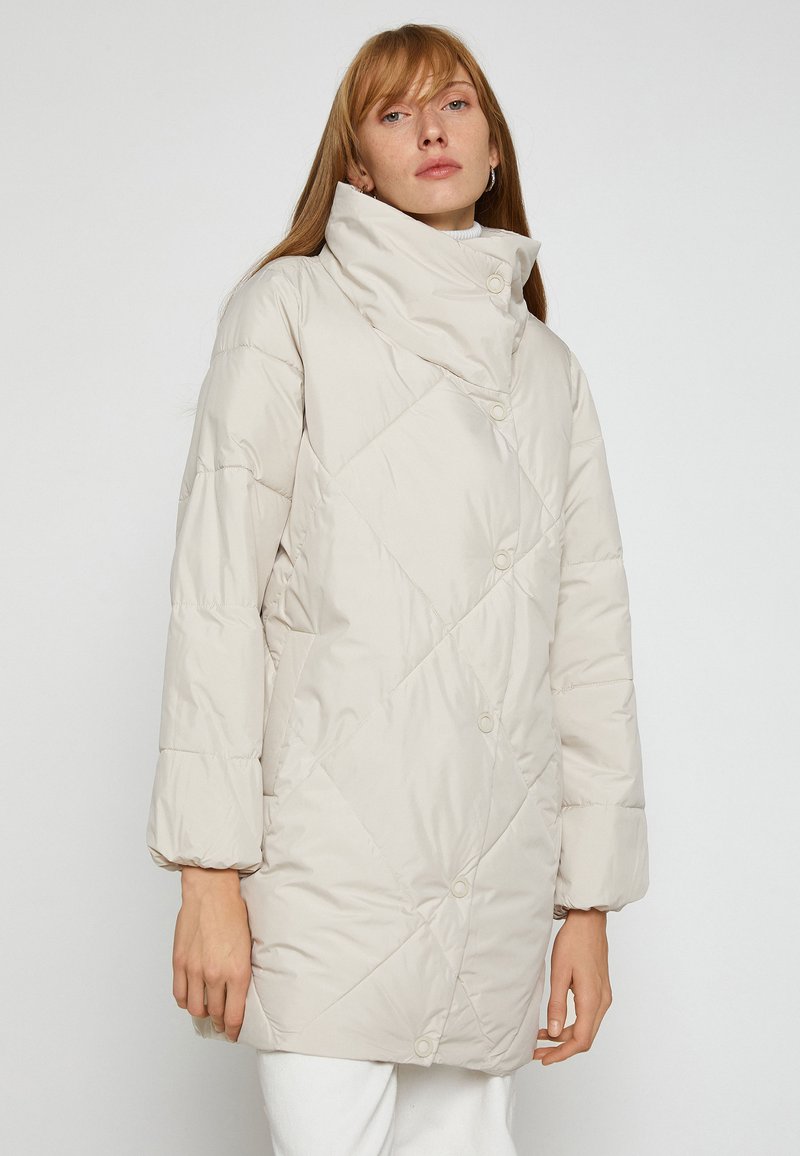 Beige quiltet puffjakke med stor krave, trykknaplukninger og skrå syninger. Har afslappede ærmer og en lang facon.