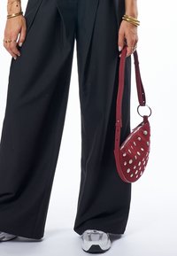 Rote Leder-Crossbody-Tasche mit silbernen Nieten, ovaler Form, verstellbarem Tragegurt, kombiniert mit schwarzen weitem Hosen und weißen Sneakers.