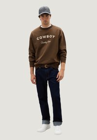 Felpa marrone con scritta "COWBOY Country Club", jeans blu scuro, sneakers bianche e cappellino grigio. Sfondo semplice.