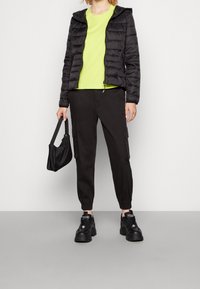 Černá prošívaná bunda s kapucí, neonově zelené tričko, černé cargo kalhoty a černé tenisky. Součástí je malá černá kabelka a elastické manžety.