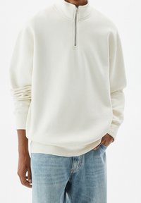 Pullover bianco in fleece con colletto a mezza zip, vestibilità rilassata, polsini a coste e orlo a bande. Abbinato a jeans azzurri chiari.