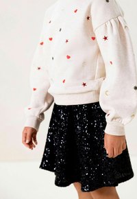 Lichtroze pullover met rode harten en gouden sterren, gecombineerd met een zwarte mini-rok van pailletten. De mouwen zijn opgeblazen en gestructureerd.