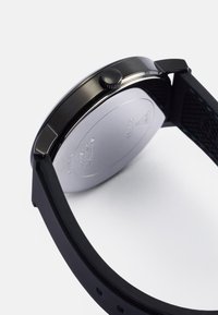 Relógio preto com um elegante estojo metálico, parte traseira prateada, bracelete em silicone texturizado e uma coroa lateral para ajuste.