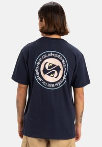 Camiseta de algodón azul marino con un gran logo circular en la parte de atrás que presenta elementos de diseño únicos y un ajuste cómodo y holgado.