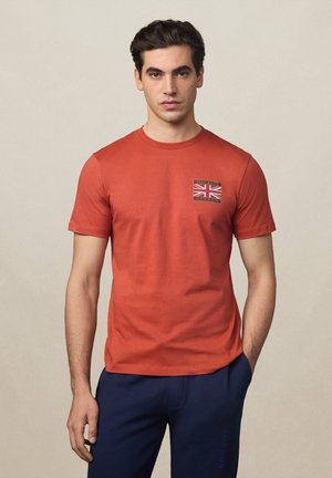 Mies, jolla on ruosteenpunainen t-paita, jossa on Union Jack -merkki ja "Hackett London" -teksti, yhdistettynä tummansinisiin housuihin, seisoo yksinkertaisen taustan edessä.