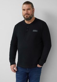 Czarna koszulka henley z długim rękawem, okrągłym dekoltem, listwą na trzy guziki i przeszywanym detalem naszywki. Noszona z niebieskimi dżinsami.