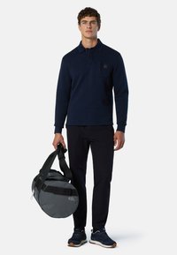 Polo de manga larga en color marino con un bolsillo en el pecho, combinado con pantalones oscuros. El modelo lleva un bolso de deporte gris con detalles en negro.