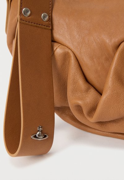 Τσάντα από ανοιχτό καφέ δέρμα με φαρδιά λαβή, ασημί πριτσίνια και μικρό ασημί λογότυπο σε σχήμα σφαίρας κοντά στον κάτω άκρο της λαβής.