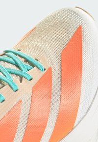 Primo piano di una scarpa da corsa beige e arancione con lacci turchesi e tomaia in tessuto testurizzato.