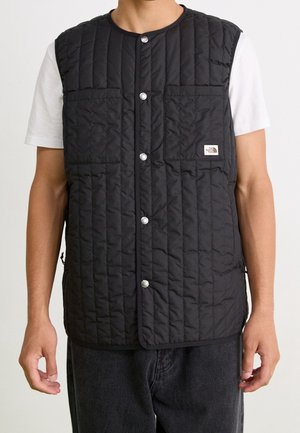 Mann trägt eine schwarze gesteppte Weste mit Druckknöpfen über einem weißen kurzärmeligen Hemd und dunklen Hosen, Markenetikett der Weste auf der Brusttasche.