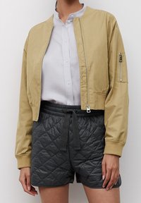 Giacca bomber tan con zip frontale, polsini a costine e tasca con zip sulla manica, abbinata a pantaloni shorts trapuntati scuri con vita regolabile tramite cordino.
