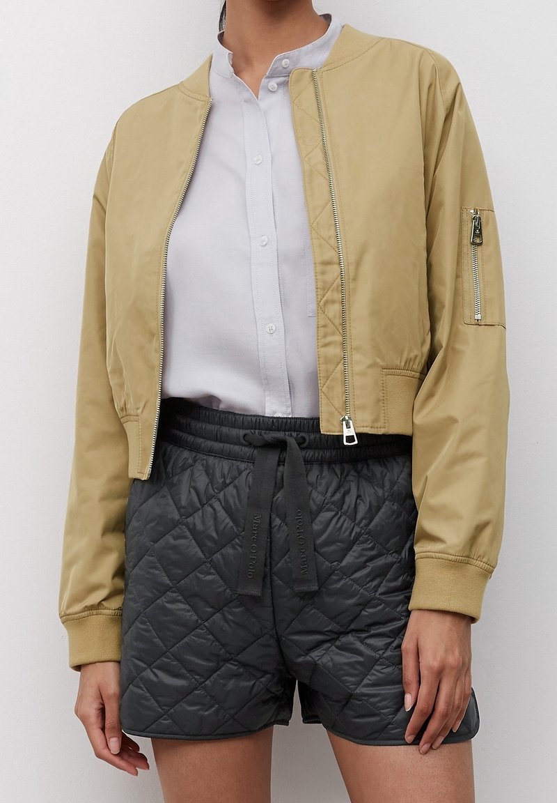 Giacca bomber tan con zip frontale, polsini a costine e tasca con zip sulla manica, abbinata a pantaloni shorts trapuntati scuri con vita regolabile tramite cordino.