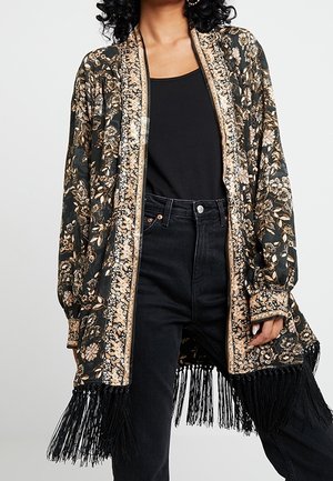 Femme portant un kimono noir à fleurs avec broderie beige et franges, par-dessus un débardeur noir et un jean noir taille haute.