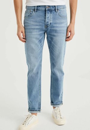 Man draagt lichtblauwe opgerolde spijkerbroek en witte sneakers, staand tegen een effen achtergrond.