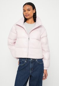 Femeie purtând o geacă matlasată roz deschis, închisă până la jumătate peste un top gri și blugi largi bleumarin, stând în picioare pe un fundal simplu.