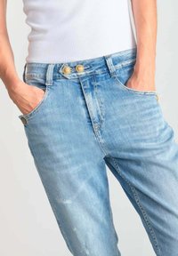 Jean en denim bleu clair avec une coupe décontractée, doté de deux boutons dorés et de poches. Le tissu présente une usure et une décoloration subtiles.