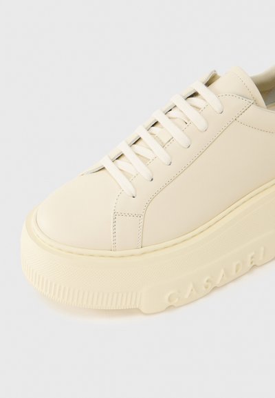 Baskets couleur crème avec tige en cuir lisse, bout rond et semelle en caoutchouc épaisse. Elles présentent des lacets ton sur ton et un logo embossé sur le côté.