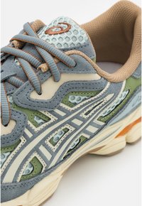 Grå och grön sneaker med nätförstärkningar, gummisula, beige detaljer och reflekterande ränder. Har texturerade material och rundad form.