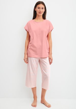 Frau steht barfuß und trägt ein lockeres, pinkfarbenes Kurzarmoberteil und weiße knöchellange Hosen mit dünnen pinkfarbenen horizontalen Streifen.