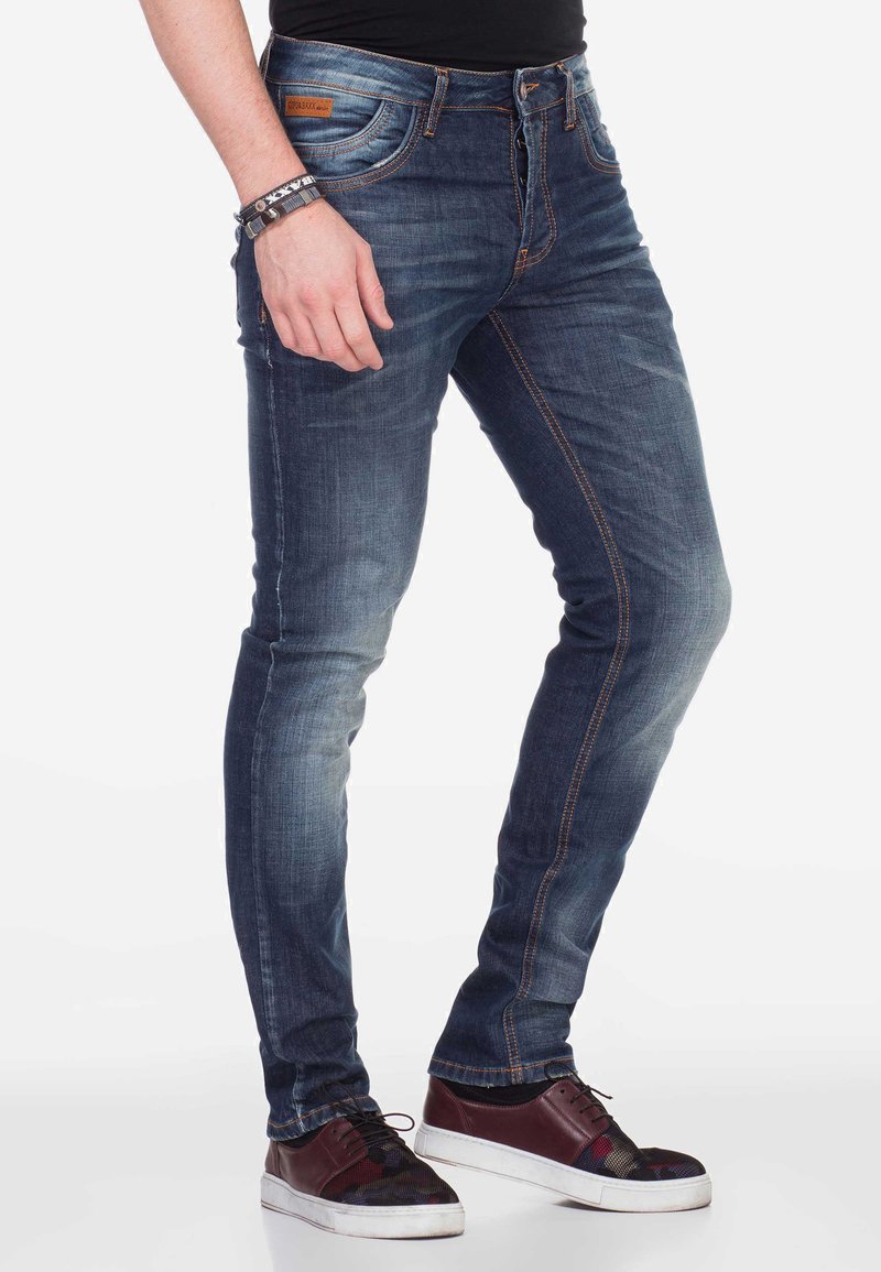 Schmale blaue Jeans aus Denim mit verblassten und abgetragenen Stellen, orangefarbener Naht und Knopfverschluss. Kombiniert mit burgunderfarbigen Sneakers mit Muster.