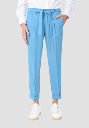 Donna che indossa pantaloni azzurri a vita alta con pieghe, cintura annodata, orli risvoltati, sneakers bianchi e camicia bianca a maniche lunghe.