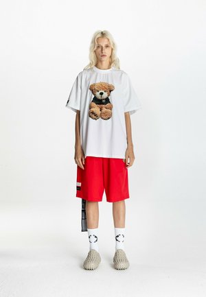 Persona rubia con una camiseta blanca de gran tamaño con un dibujo de un osito, pantalones cortos rojos hasta la rodilla, calcetines blancos y zapatos beige con textura.