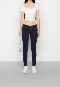Top corto blanco con flores, pantalones ajustados azul marino y zapatillas blancas de plataforma. Incluye un bolso cruzado azul. Diseño simple y ajustado con textura suave.