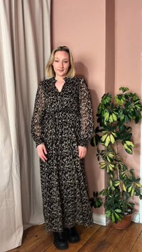 Eine Frau steht drinnen und trägt ein langes schwarz-beiges Kleid mit Muster und transparenten Ärmeln, schwarze Stiefel, neben einer grünen Topfpflanze und einer pfirsichfarbenen Wand.