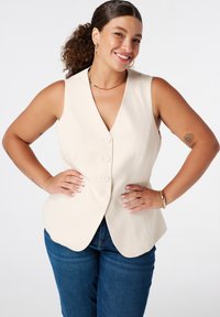 Beige mouwloos vest met een V-hals, vijf knopen aan de voorkant en een getailleerde pasvorm, gecombineerd met blauwe jeans. Glad stofmateriaal.