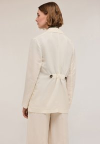 Blazer beige chiaro in lino, con schiena arricciata e bottoni neri, colletto a revers e maniche lunghe. Tessuto liscio e vestibilità sartoriale.