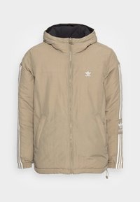 Beige puffjacka med svart inredning. Har en framzipper, två sidofickor och tre vita ränder längs ärmarna. Adidas-logotypen är närvarande.