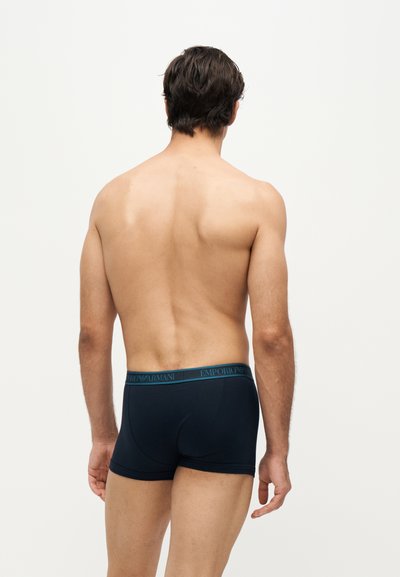 Homme debout, dos face à l'avant, portant un boxer bleu marine Emporio Armani sur un fond uni.