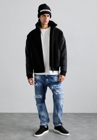 Veste zippée noire avec une texture en polaire et des manches contrastées, associée à un jean bleu déchiré et des baskets noires. T-shirt blanc en dessous.