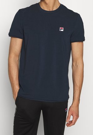 Mann trägt ein marineblaues T-Shirt mit kurzen Ärmeln und kleinem Fila-Logo auf der Brust, kombiniert mit einer schwarzen Hose, steht vor einem schlichten Hintergrund.