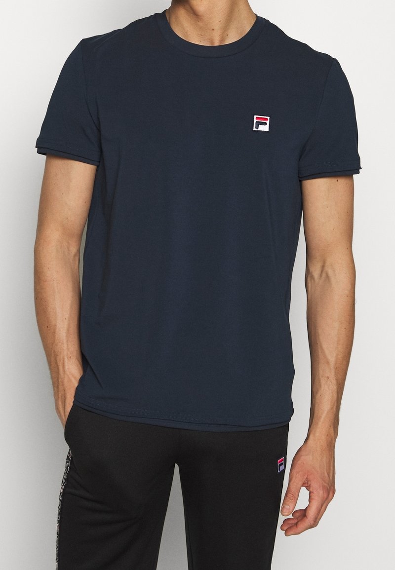 Homme portant un t-shirt bleu marine à manches courtes avec un petit logo Fila sur la poitrine, associé à un pantalon noir, debout devant un fond uni.