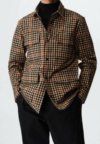 Chemise à carreaux marron et noire avec deux poches avant, fermeture à boutons, et texture douce. Portée par-dessus un col roulé noir.