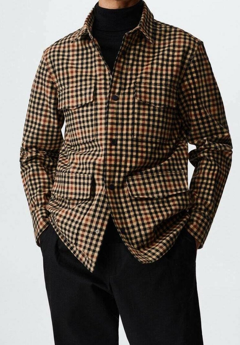 Chemise à carreaux marron et noire avec deux poches avant, fermeture à boutons, et texture douce. Portée par-dessus un col roulé noir.