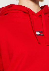 Sweat à capuche rouge avec cordons et petit patch logo Tommy Jeans sur la poitrine, présenté du cou au haut du torse.