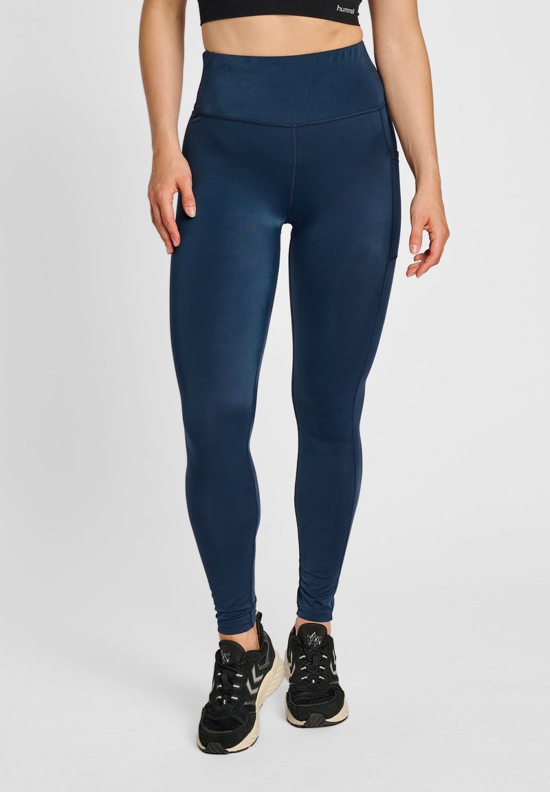 Höga marinblå leggings med en slät yta, med en sidoficka och förstärkta sömmar, tillsammans med svarta träningsskor.