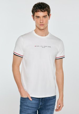 Młody mężczyzna z krótkimi kręconymi włosami, ubraną białą koszulkę z czerwono-czarnymi paskowanymi krawędziami rękawów, tekstem na piersi i niebieskimi dżinsami.