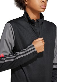 Schwarze Zip-up-Jacke mit grauen Ärmeln, die drei horizontale Streifen an den Bündchen und einen hohen Kragen aufweist. Hergestellt aus strukturiertem Stoff.