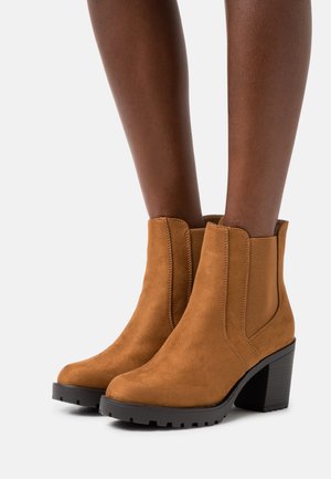 Botas de tobillo de ante color cuero con paneles laterales elásticos, punta redondeada y suela de goma negra texturizada; cuentan con un tacón grueso para ofrecer soporte.