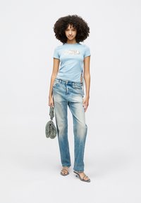 Camiseta ajustada azul-claro com um logo metálico, combinada com jeans de perna reta desbotados, segurando uma bolsa verde texturizada e usando sandálias com tiras.